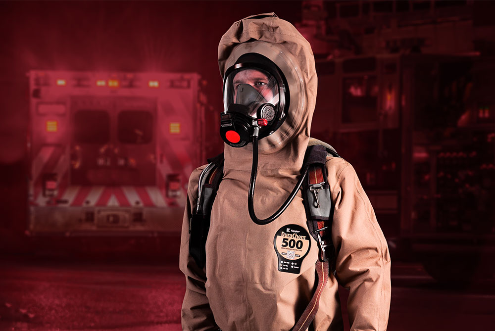 Responder in a tan Kappler DuraChem 500 suit and respirator beside an ambulance