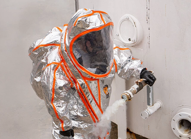 Responder in a silver and orange aluminized Kappler suit working a leaking valve amid vapor